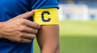© ManuStockStudio - Close up of a soccer team captain's arm as they adjust their yellow armband with a black letter C during a match on a green field stadium with goal in background