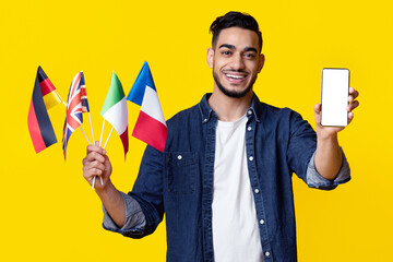 Happy arab man showing smartphone with empty screen and bunch of international flags, yellow studio background, panorama, mockup. Foreign language learning mobile application concept
