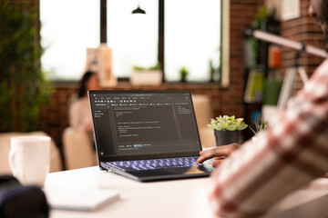 African american man sitting at desk, coding on laptop and troubleshooting website scripts. Young male IT specialist working remotely on web development from brick wall apartment.