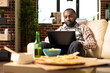 © DC Studio - African american man seated on couch with serious expression, working on laptop during downtime. Male entrepreneur surrounded by snacks and drink, completes company tasks in brick wall home.