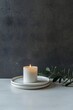 © Imagine  - Minimalist white candle burning on ceramic plates with green leaves on marble surface against dark textured wall, perfect for relaxation, meditation, spa, and home decor branding