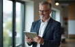 © liang - Middle aged happy business man professional manager executive standing in office using tablet looking away. Busy smiling mature 50 years old businessman investor thinking of future. Copy space.