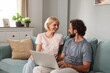 © Stockphotodirectors - In a serene living room, a couple shares a cheerful moment as they engage with a laptop on a comfortable couch. Natural light brightens the space, creating an inviting atmosphere.
