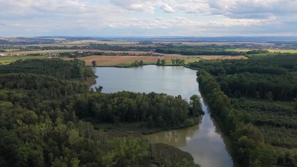 Wall Mural - drone view of a pond surrounded by fields and meadows in summer, with a nearby village under cloudy 