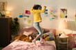 © pressmaster - Caucasian girl child wearing headphones jumping on bed in decorated bedroom surrounded by stuffed animals and colorful drawings on wall, expressing energetic playful mood