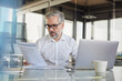 © Westend61 - Businessman reading document at modern office desk with laptop