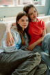 © m-art - Two young women sit closely together on a couch, sharing a cozy moment. They are dressed casually, reflecting the comfort of a relaxed afternoon spent indoors.