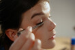 © m-art - A woman sits at a wooden table applying mascara to her eyelashes. Natural light fills the room, highlighting her focused expression and delicate features. She is immersed in her makeup routine.