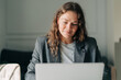 © Ilona - A middle-aged woman sitting at home in a business suit works on a laptop.