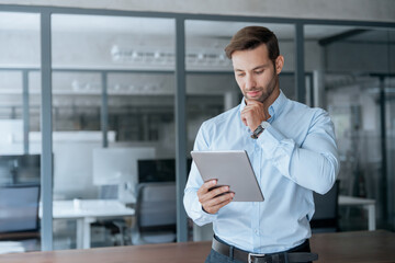 Focused Latin entrepreneur businessman holding digital pc tablet standing in office. Smiling European young man working on trade online using touchpad fintech computer for business work. Copy space