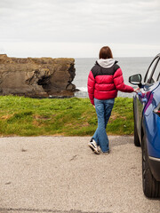 Wall Mural - A woman in a red jacket stands next to a blue car in a parking lot and looking at a stunning nature scene with cliff, rough stone coastline and cloudy sky. Travel and tourism. West of Ireland.