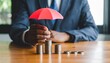 © photostockatinat - A businessman uses a red umbrella to protect a stack of coins, symbolizing financial security and protection