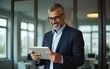 © Susan - Focused senior mature Indian or Latin entrepreneur businessman holding digital pc tablet standing in office. Smiling hispanic man in suit working using touchpad computer for business work project