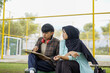 © Fajar - Two students studying outdoors together, discussing a tablet. A young man with headphones points at the screen, while a woman in a hijab attentively looks on. Collaboration and learning concept