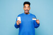 © ViDi Studio - Young surprised man wear sweatshirt casual clothes using mobile cell phone hold credit bank card doing online shopping order delivery booking tour isolated on plain blue background. Lifestyle concept.