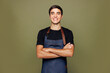 © ViDi Studio - Young man barista bartender barman employee wear jeans apron black t-shirt work in coffee shop hold hands crossed folded look camera isolated on plain green background. Small business startup concept.