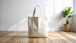 © RatihMeiriga - A blank white canvas tote bag stands on a wooden floor, with a potted plant and sunlight in the background.