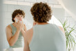 © m-art - A young man stands in front of a mirror applying shaving cream to his face. The bright bathroom features greenery and a modern design, creating a refreshing atmosphere for personal care.