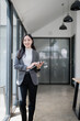 © MINAE - Young Asian businesswoman smiling while walking through a modern office corridor, using a digital tablet amidst bright glass walls, embodying professionalism and confidence