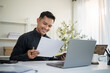 © matinee - Smiling businessman reading documents while working on laptop in modern office.