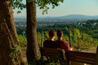 © Klym Bykov e.U. - Horizontal photo of a couple sitting on a bench among trees, looking at vineyards and hills. Warm autumn sunlight bathes the scene, creating cozy, romantic, and rustic atmosphere.