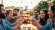 © fathiqi - A group of happy young friends in denim shirts toasting with glasses of beer at an outdoor patio during sunset.