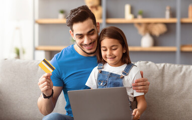 Portrait of smiling father and kid shopping together on Internet, using laptop and credit card, sitting and hugging on couch at home, looking at computer screen and smiling, order food, copy space