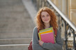 © noiteterna - Persona pelirroja con mochila y suéter gris sostiene cuadernos coloridos en escalinata de piedra con barandilla decorativa. Sonríe directamente a cámara en entorno universitario clásico y acogedor.