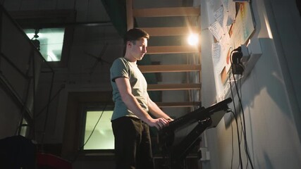 Young technician stands in dim backstage room, staring at illuminated control panel under desk lamp near cork board filled with handwritten notes. Scene captures quiet focus in industrial workspace