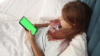Child girl using smartphone looks at green screen, presses button to view chromakey online lying on bed.
