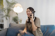 © kamiphotos - A cheerful young woman sits casually on a sofa, holding a smartphone to her ear while talking and smiling. With headphones around her neck, she looks relaxed, expressive, and engaged.