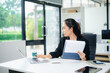 © laddawan - Smiling Asian businesswoman texting on phone at modern office desk with laptop, coffee, and documents