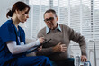 © NanSan - A young Caucasian nurse provides medical consultation to an elderly Caucasian man sitting on a sofa, addressing chronic conditions like obesity, hypertension, diabetes,heart disease, dementia, joint