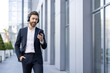 © Liubomir - Businessman in a stylish suit, wearing headphones and walking confidently near a modern office building, engaging with a smartphone app while enjoying a sunny day in the city