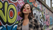 © Shantuna - A young woman with sunglasses, a jacket and red lipstick is standing in front of a graffiti wall looking up with a confident expression