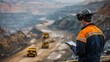 © Justlight - Mining professional wearing reflective safety gear inspects mining progress on a handheld tablet with sprawling openpit terraced benches and heavy duty haul trucks crawling below.