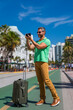A traveler with camera taking photo on streets of Miami. A man traveler enjoys his summer vacation in a tropical city. A traveler tourist with suitcase on summer city. Man traveler walk on city.