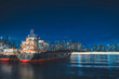 A cargo ship is docked by the city waterfront at night, with the city skyline and water reflection visible.