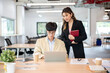© bongkarn - Asian businesswoman looking at laptop supervising an intern coworker at wooden table in the office.