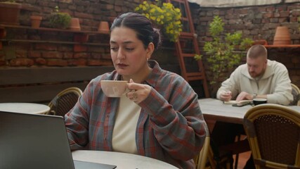 Wall Mural - Medium long shot of female multiethnic freelancer drinking coffee while working on wireless laptop at table, male Caucasian colleague taking notes in background in outdoor cafe