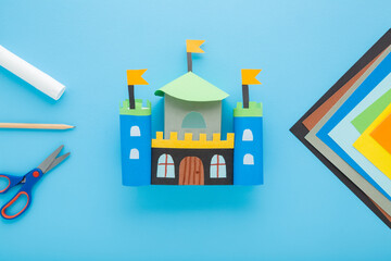 Wall Mural - Created colorful castle with towers from paper. Scissors, glue stick, pencil and application paper on light blue table background. Pastel color. Closeup. Child handcraft. Top down view.