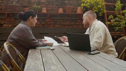 Wall Mural - Medium long shot of two diverse gen Z coworkers drinking coffee discussing tasks at table with laptop and business papers in outdoor cafe