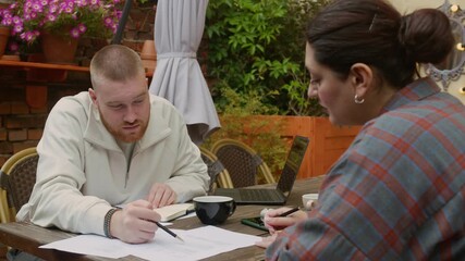 Wall Mural - Medium shot of two diverse gen Z businesspeople discussing documents at table with coffee and laptop in outdoor cafe