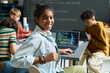 © pressmaster - Portrait of Black teenage girl smiling at camera while sitting at desk with open laptop displaying code, multiethnic teenagers working together on computers in classroom background