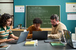 © pressmaster - Caucasian man assisting Black teenager and Caucasian teenage girl working on laptops in classroom setting, chalkboard with AI prompting instructions visible in background