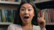 © Yuliia - Female portrait smiling happy asian woman girl student 20s looking at camera in university library showing thumbs up like gesture approval symbol successful feedback recommendation agree great result