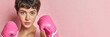 © Olena - A young female boxer with short dark hair wearing pink boxing gloves on a pink background with copy space, a banner symbolizing struggle, strength of spirit, and courage