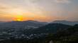 A scenic view of a town nestled among lush green mountains at sunset, with a temple structure perched on the hillside and a vibrant orange sky creating a warm, tranquil atmosphere.