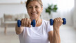 © anandart - Home training concept. Strong senior woman doing exercises with dumbbells indoors. Cheerful mature lady working out her arm muscles, keeping fit, leading healthy lifestyle during covid-19 isolation