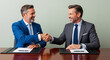 © DAYA-DAKSH - Two smiling businessmen in suits confidently shaking hands at a polished desk, sealing a successful deal with a warm handshake.